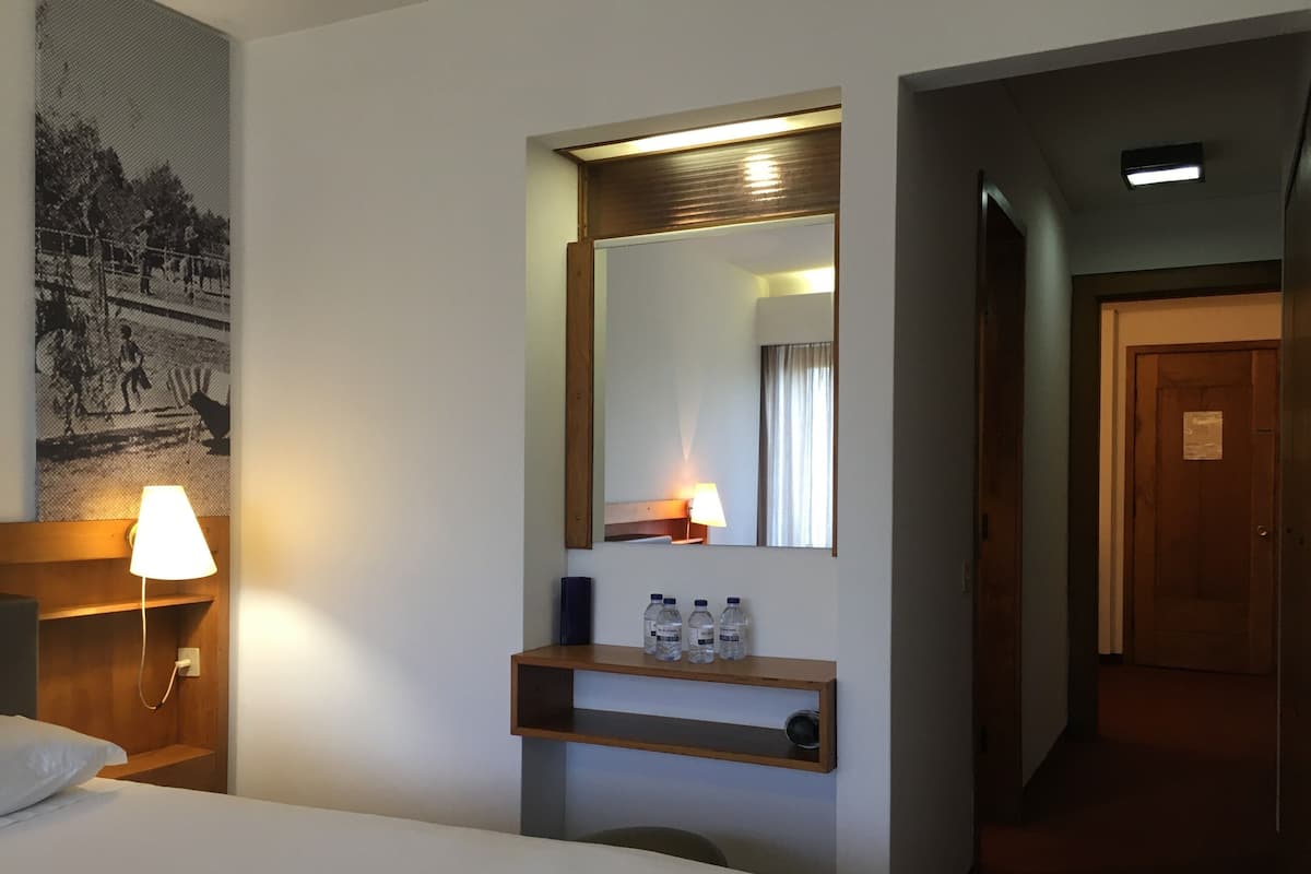 A hotel room with a bed, a nightstand with a lamp, a mirror, a shelf with water bottles, and a hallway leading to another room.