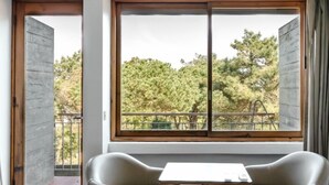 A room with a bed, two leather chairs, a small table, and a large window with a view of trees and a fence. - Parque do Rio Ofir Hotel (Esposende)