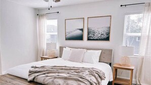 A bedroom with a large bed, a wooden bench, two bedside tables, and a window with curtains. - Coastal Getaway With Bikes & NEW Saltwater Pool (Sarasota)