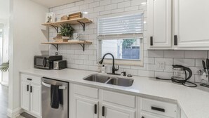 A modern kitchen with white cabinetry, a double sink, a microwave, a coffee maker, and a window with blinds. - Coastal Getaway With Bikes & NEW Saltwater Pool (Sarasota)