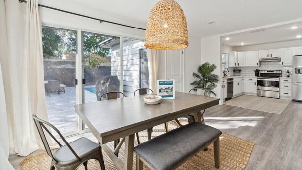 A modern kitchen with white cabinets and stainless steel appliances, a dining area with a wooden table and chairs, and a large window overlooking a patio. - Coastal Getaway With Bikes & NEW Saltwater Pool (Sarasota)