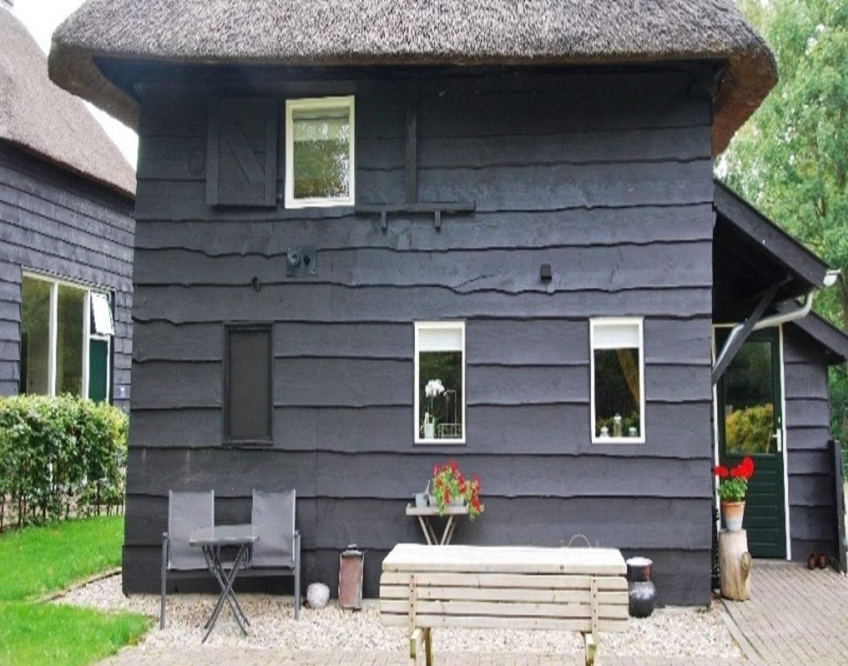 Une maison en bois peinte en noir, avec un toit de chaume, un petit espace extérieur aménagé avec un banc et des chaises, et une plante en pot.