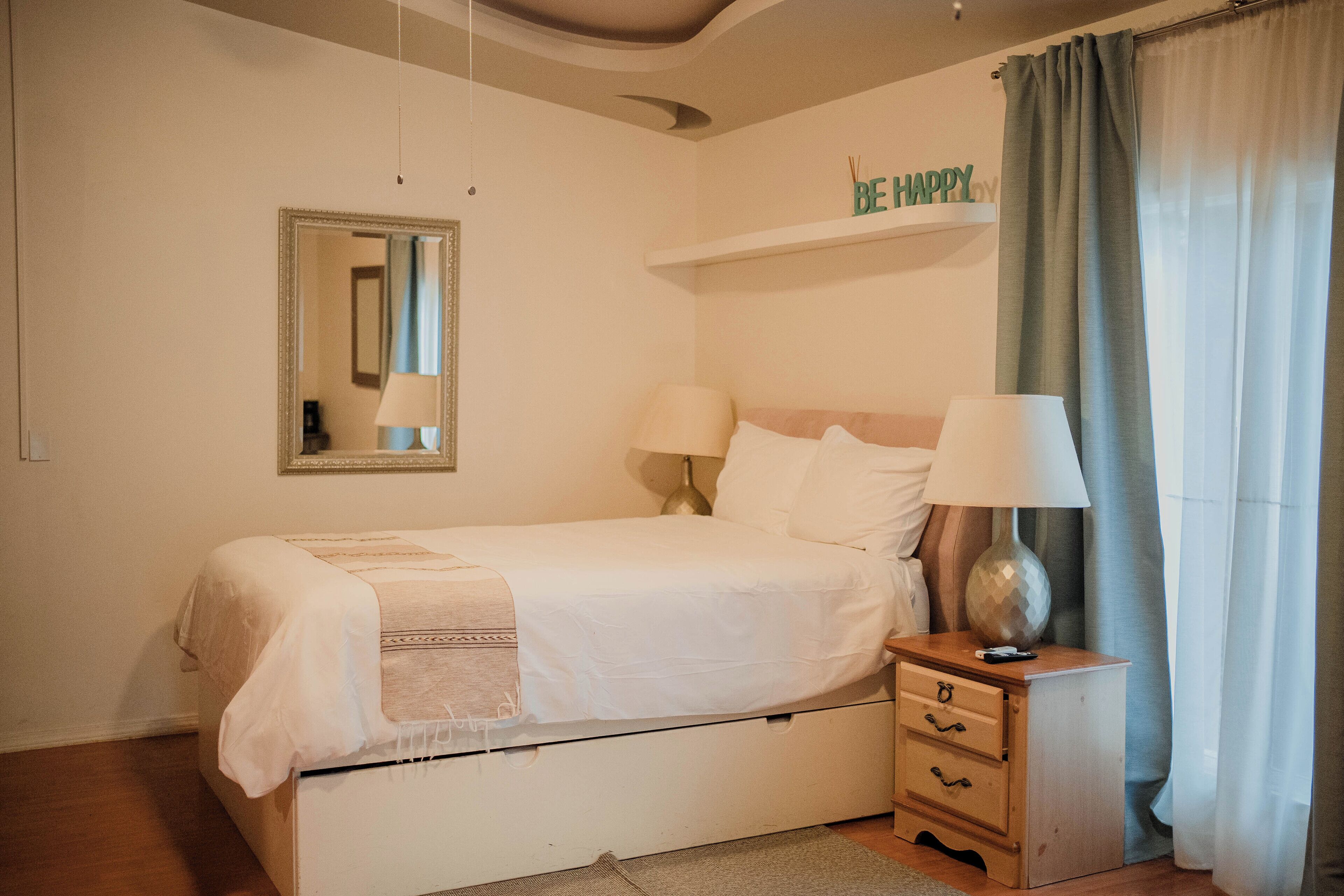 A neatly made bed with a bedside table, a mirror, and a shelf with a 'BE HAPPY' sign.