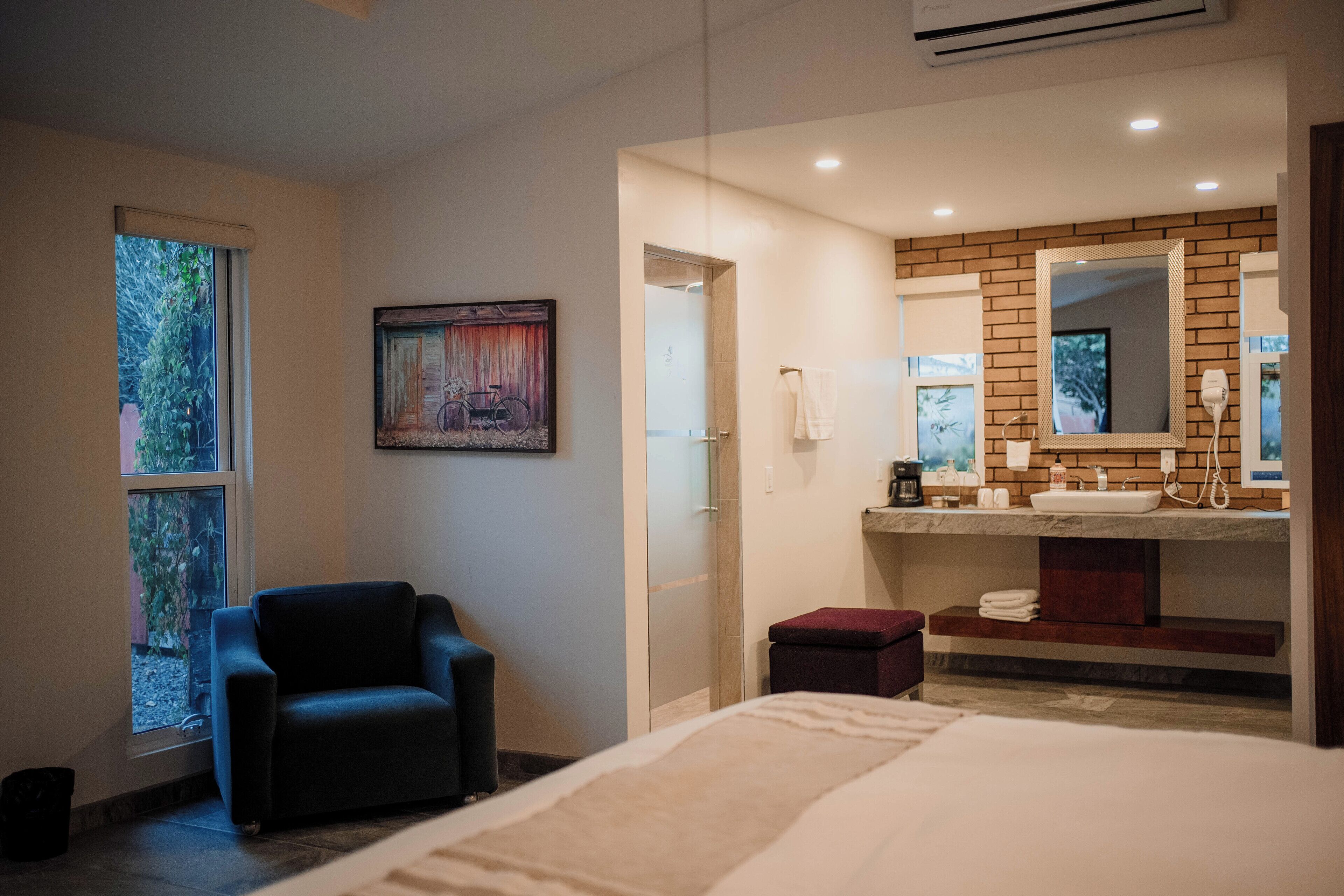 A modern hotel room with a bed, a seating area, a bathroom with a large mirror, and a painting on the wall.