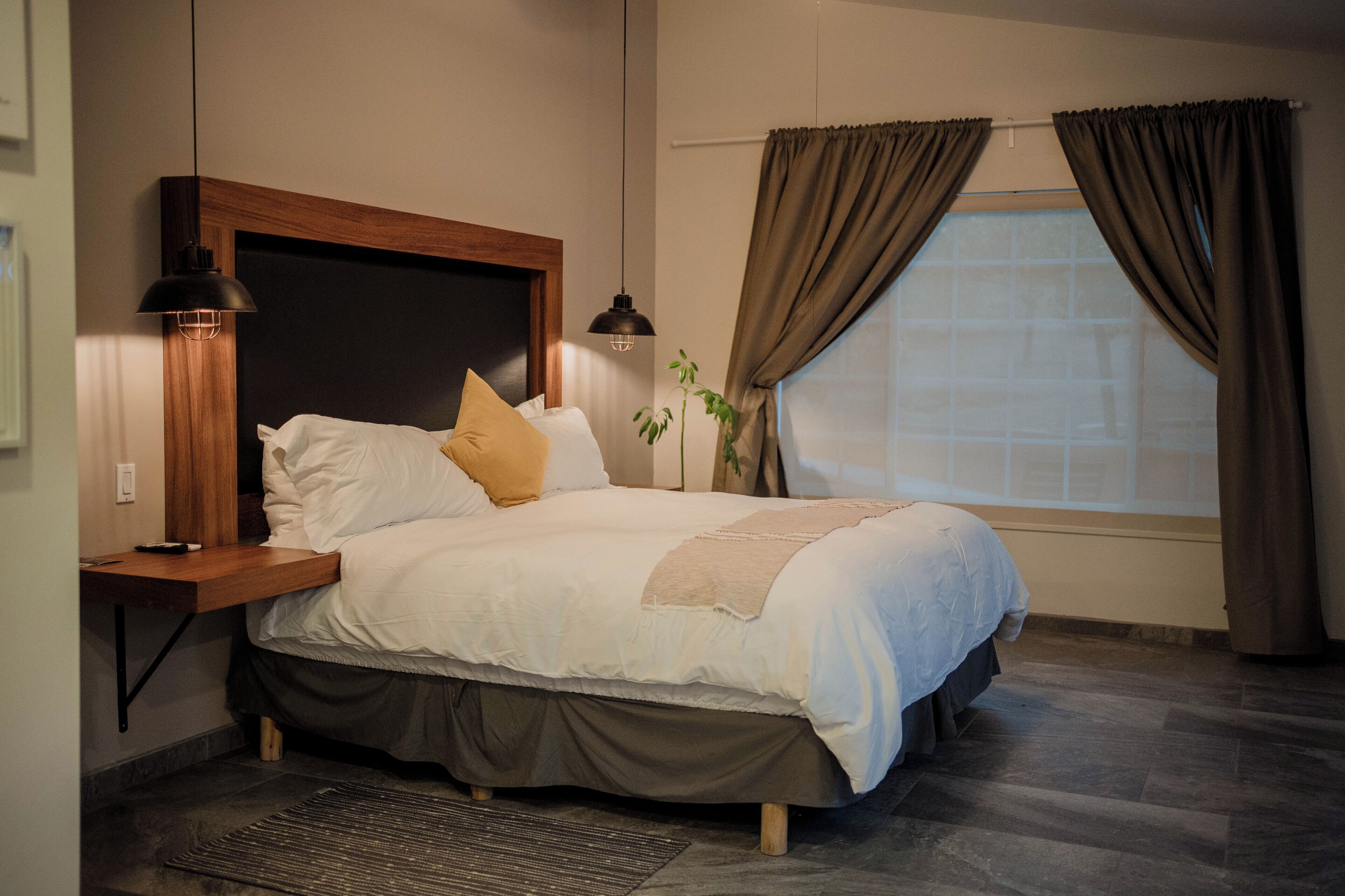 Una cama bien hecha con cabecera de madera, una mesita de noche con lámpara y una ventana con cortinas.