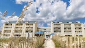 A beachfront hotel with a sandy pathway leading to the entrance, surrounded by tall grasses and a clear blue sky. - Beautifully Decorated Beachfront Condo! (Orange Beach)