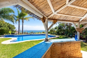 A pool area with a thatched roof, palm trees, and a view of the ocean.