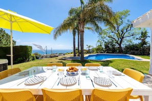 An outdoor dining area with a pool, palm trees, and a view of the ocean.
