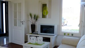 A living room with a white sofa, a small white table with decorative items, a television on a white stand, and a potted plant. - Heathside Family Retreat (Wietzendorf)