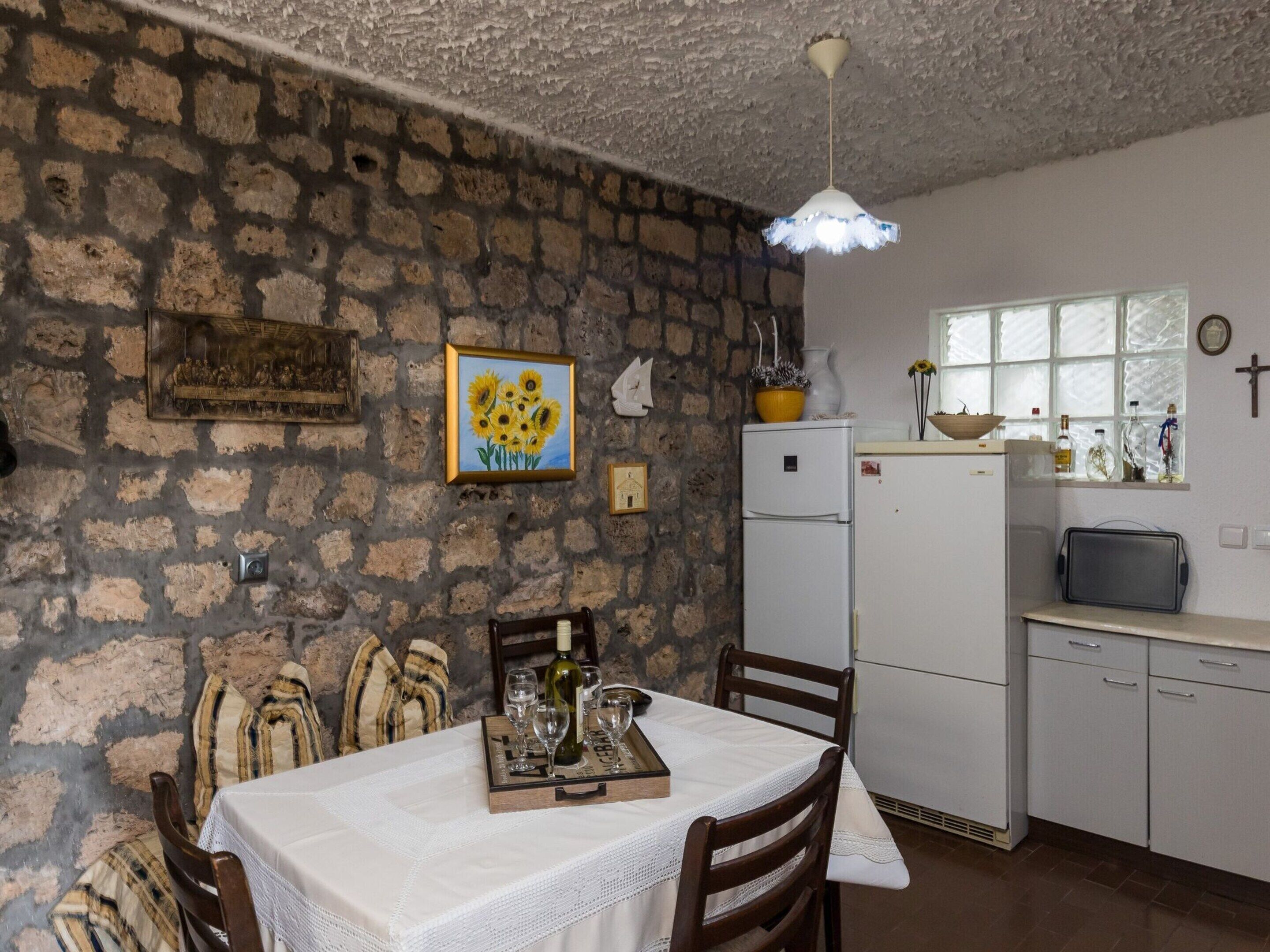A dining area with a stone wall, a white table with a lace tablecloth, wooden chairs, and a tray with wine glasses and a bottle.