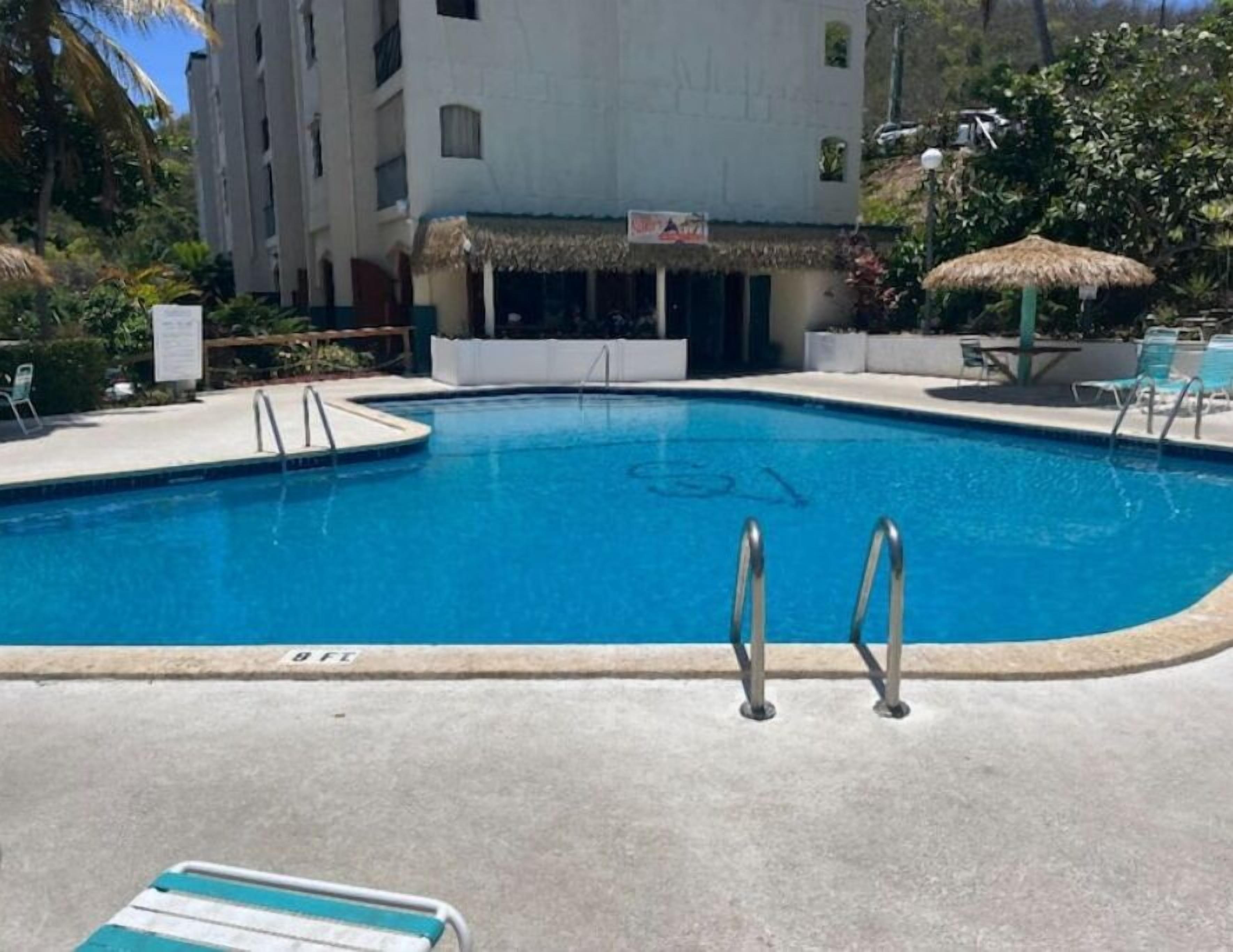 Une piscine avec deux échelles, des chaises longues et, au fond, un bâtiment doté d’un toit de chaume.