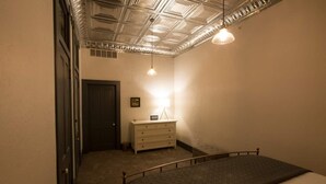 Une petite pièce faiblement éclairée, avec un lit simple, une commode et une porte.