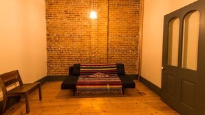 Une pièce chaleureuse avec un mur en briques, un plancher en bois, un canapé noir agrémenté d’une couverture colorée et une chaise en bois.