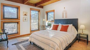 A bedroom with a large bed, wooden beams on the ceiling, multiple windows, and a view of trees. - Our sunny, solar-powered home is located on the biking path that leads to Salida’s quaint downtown. The house has a rural feel and fantastic panoramic views, but is just 3 miles from all the restaurants, galleries, shops, and parks of Salida. (Salida)