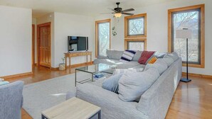 A modern living room with a grey sofa, a glass coffee table, a wooden side table, a ceiling fan, and large windows. - Our sunny, solar-powered home is located on the biking path that leads to Salida’s quaint downtown. The house has a rural feel and fantastic panoramic views, but is just 3 miles from all the restaurants, galleries, shops, and parks of Salida. (Salida)