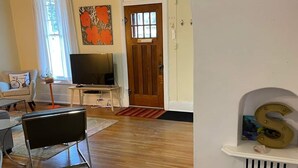 Una sala de estar con piso de madera, televisor, puerta y un respiradero en la pared.