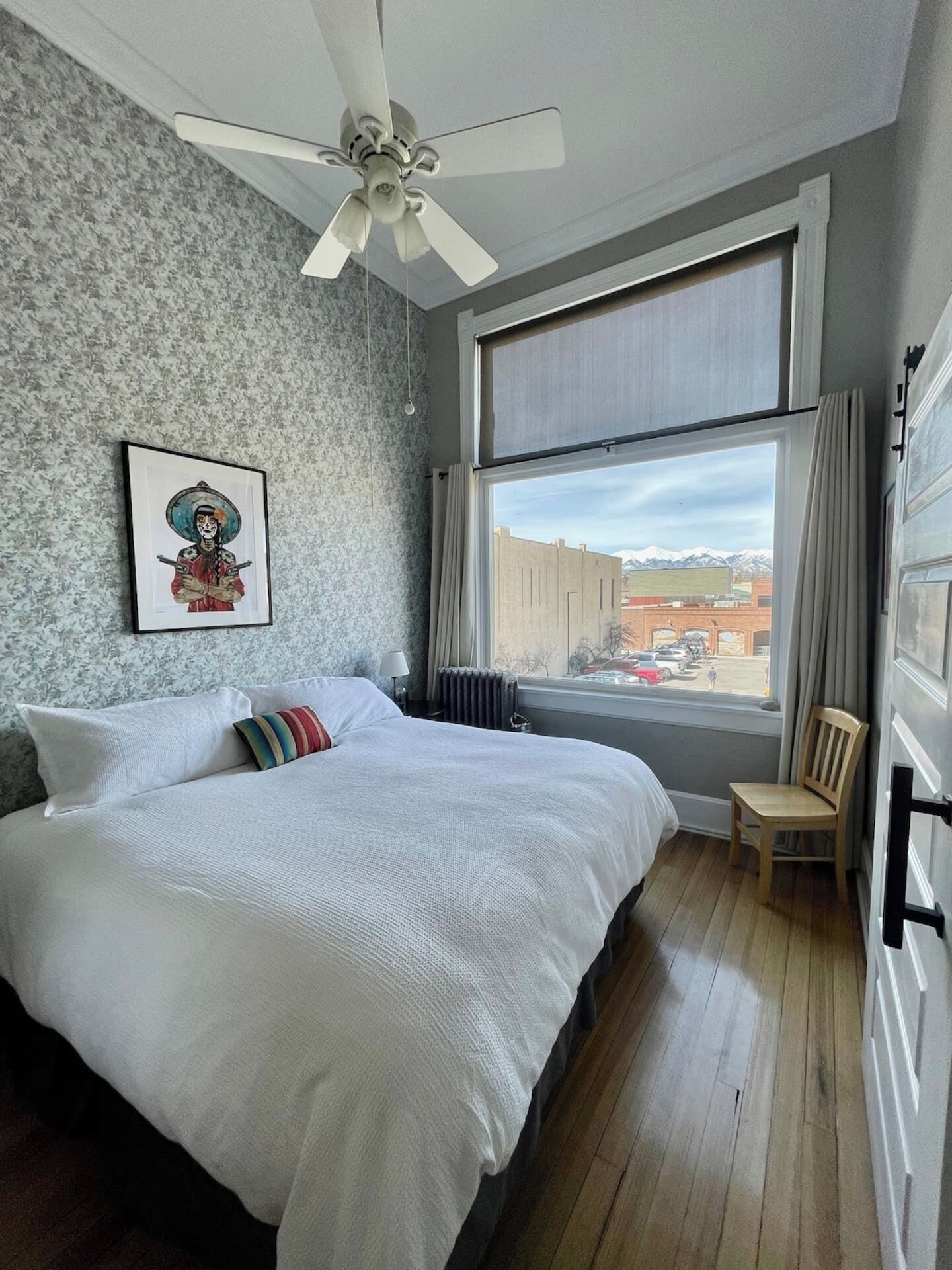 Een slaapkamer met een groot bed, een plafondventilator, een raam met uitzicht op gebouwen en bergen, en een ingelijst kunstwerk aan de muur.