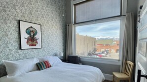 A bedroom with a large bed, a ceiling fan, a window with a view of buildings and mountains, and a framed picture on the wall. - 2nd story condo overlooking the F Street Pedestrian mall, downtown Salida! (Salida)
