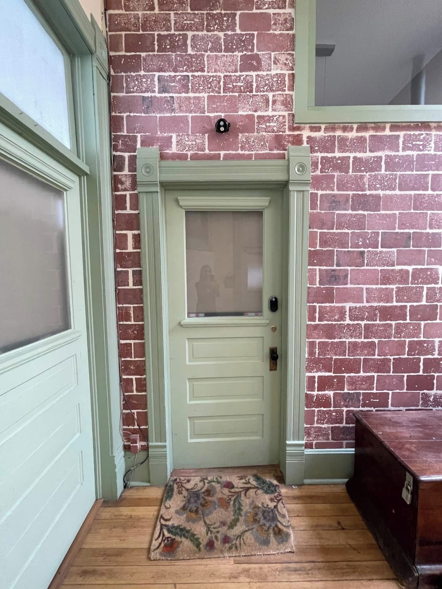 A door with a frosted glass panel, a small rug with floral patterns, and a brick wall with a green trim.