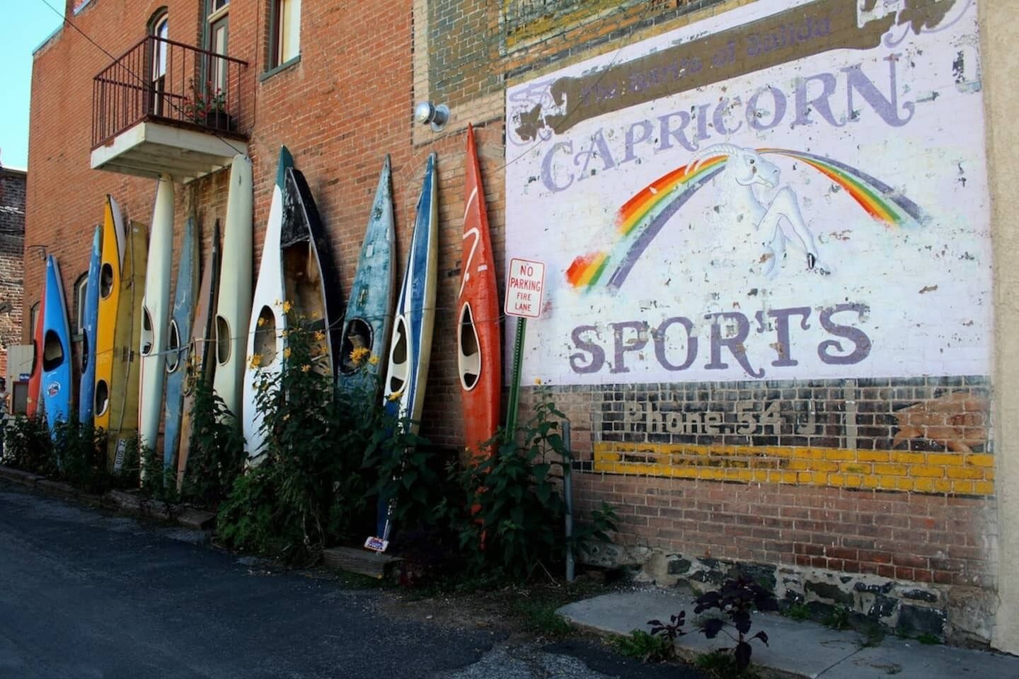 Un edificio con un mural publicitario de 'Capricorn Sports' y varias kayaks alineadas contra la pared.