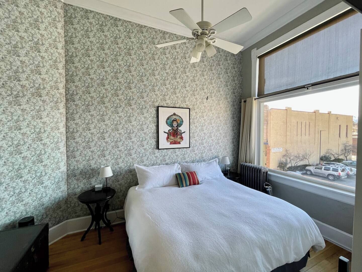 A bedroom with a bed, a ceiling fan, a window with a view of a building, and a wall with a floral pattern.