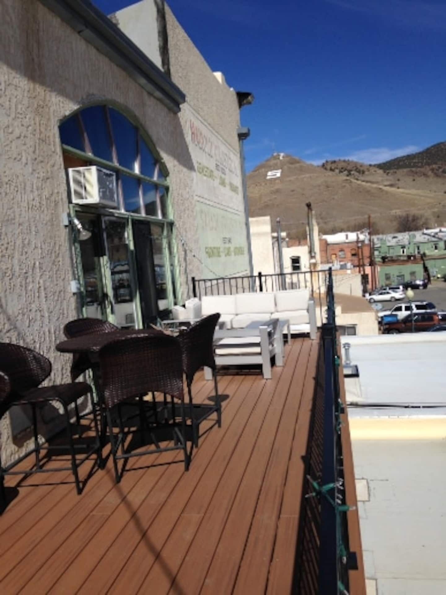 A wooden deck with outdoor seating, a building with signage, and a parking area in the background.