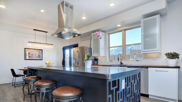 A modern kitchen with a central island, bar stools, stainless steel appliances, and a large window providing a view of the cityscape. - 2 story condo w/ balconies, hovering above the action of the downtown scene. (Salida)