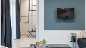 A modern living room with a flat-screen TV mounted on a teal wall, a black sofa, a small side table with a plant, and a glass coffee table. - 85 sqm  in the Heart of Athens w/Rooftop Deck (Athens)