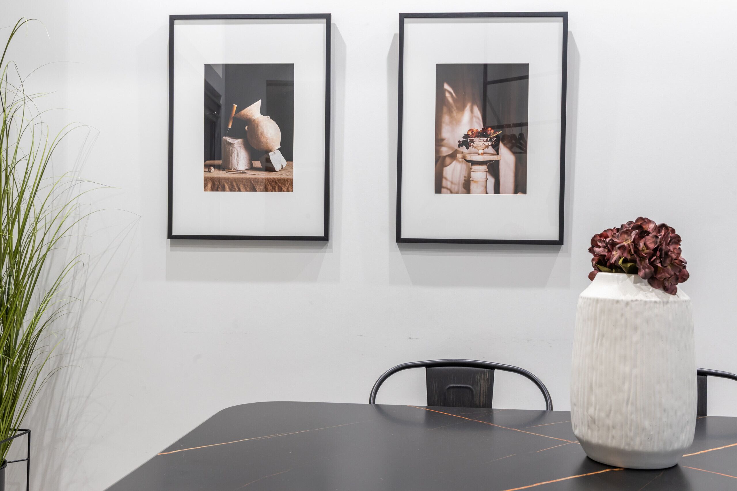 A dining table with two framed pictures on the wall, a white vase with red flowers, and a potted plant.