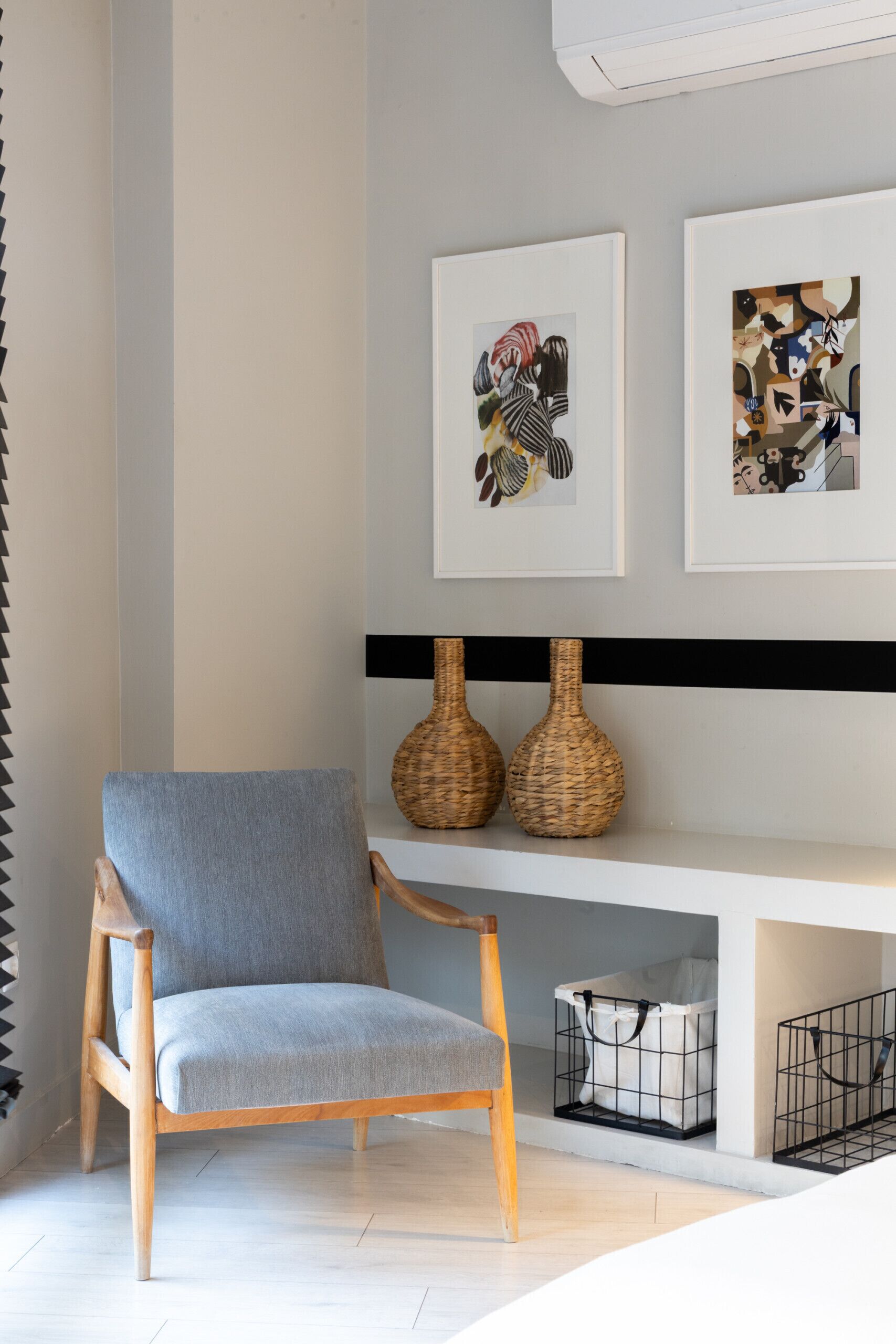 Un salon moderne avec un fauteuil gris, deux vases en osier posés sur une étagère et un tableau abstrait encadré au-dessus.