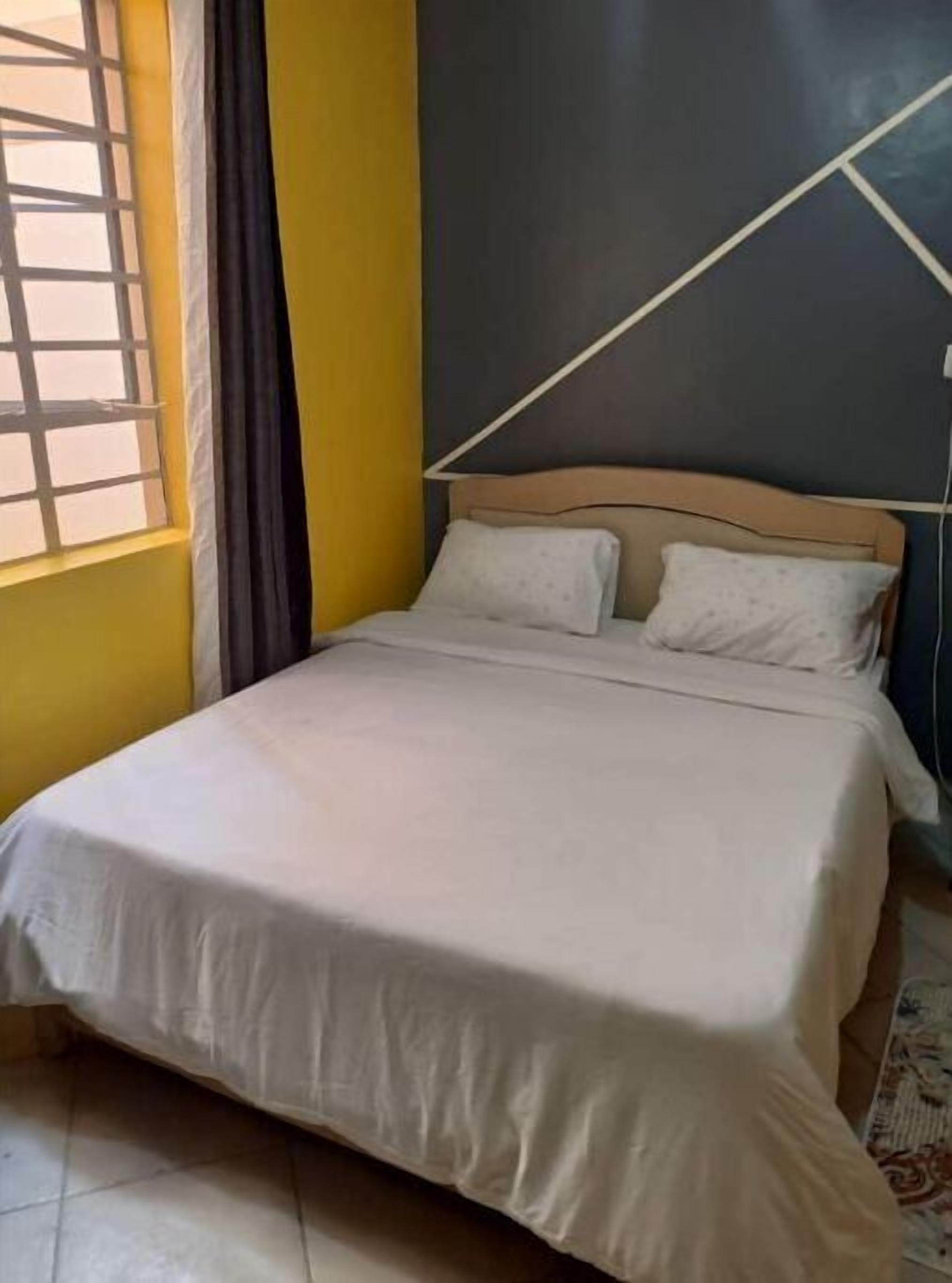 Ein Bett in einem Zimmer mit gelben Wänden, einem Fenster mit Jalousien und einem schlichten Kopfteil.