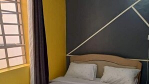Un lit dans une chambre aux murs jaunes, une fenêtre avec des stores et une tête de lit sobre.