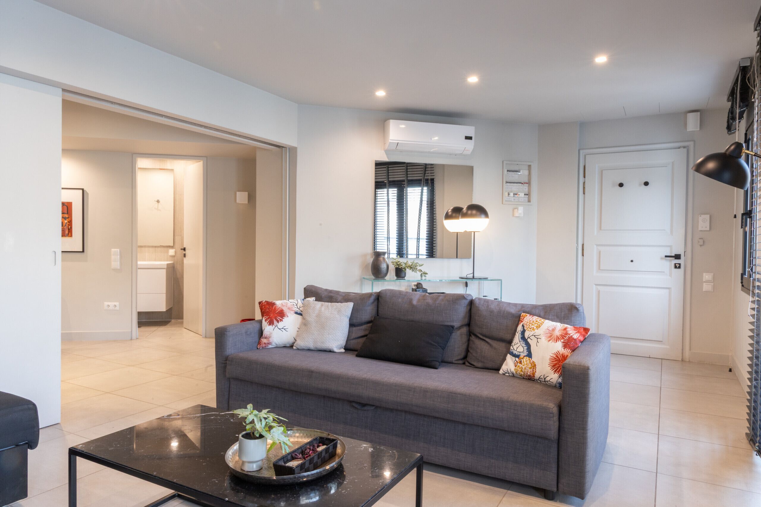 A modern living room with a grey sofa, a black coffee table, and a small plant. There is a white door and an air conditioning unit on the wall.