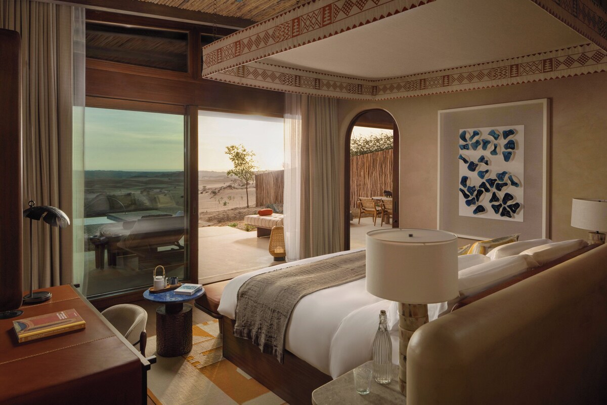 A hotel room with a large bed, a desk, a chair, a table, and a view of the desert landscape.