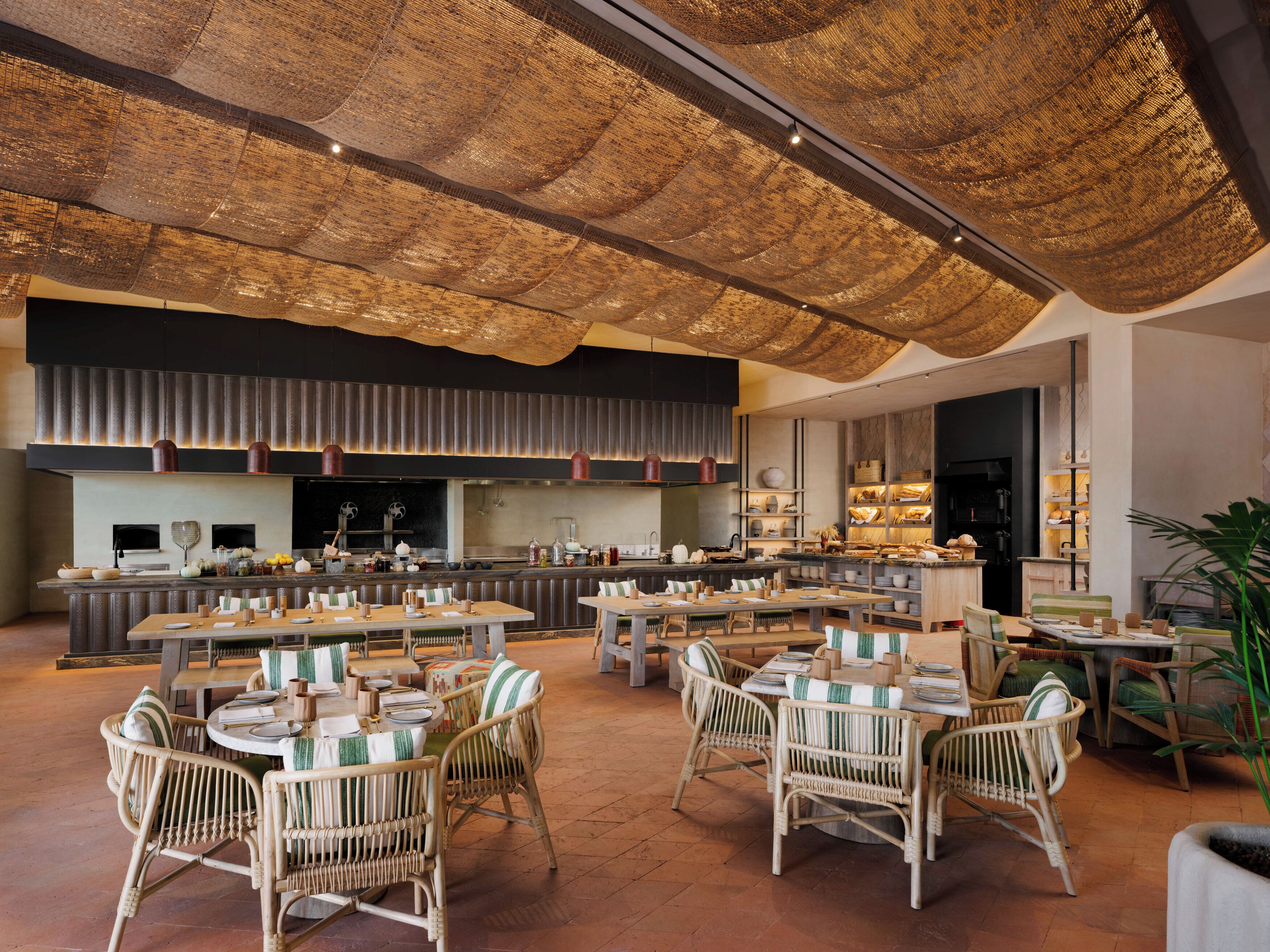 Een ruim restaurant met hoge plafonds, rieten meubels en een moderne keuken op de achtergrond.