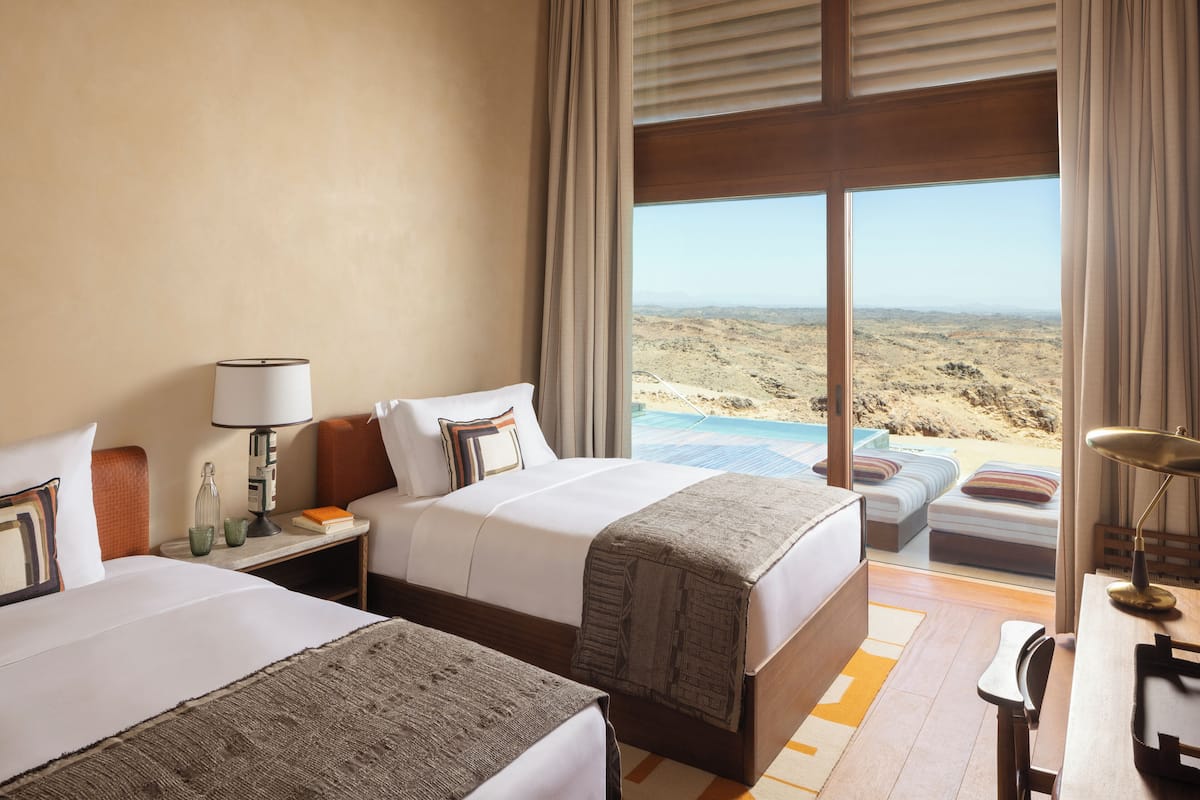 Relax in a serene space with two beds, a desk, and a nightstand, offering a view of the desert landscape and pool area.