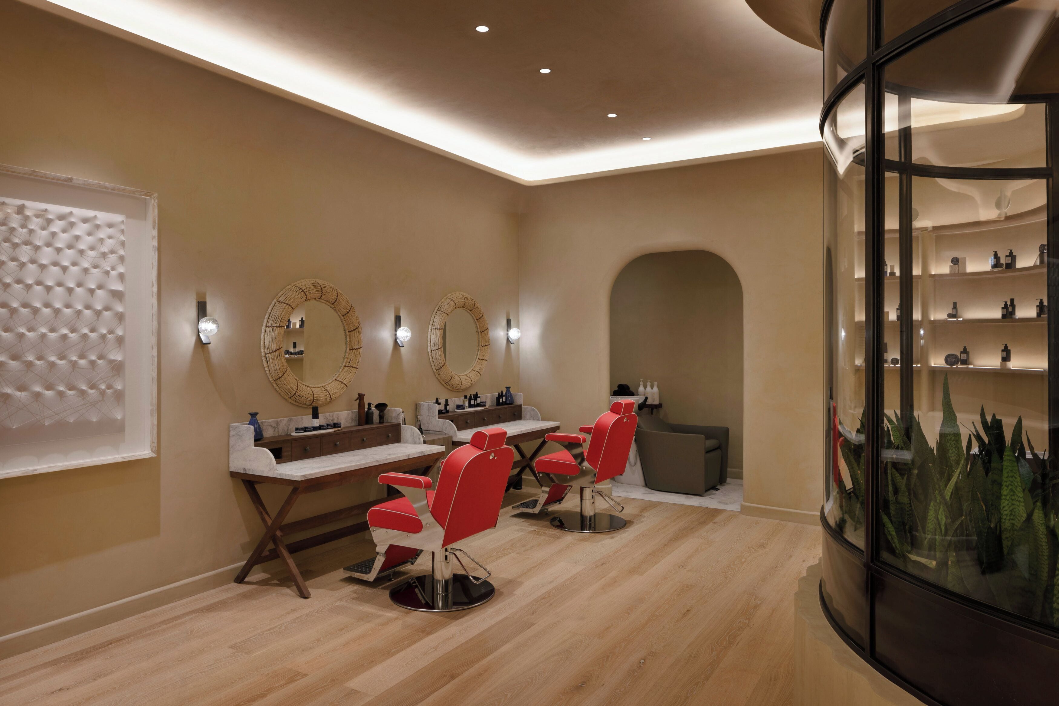 A salon with two red chairs, a mirror, and a shelf with toiletries.