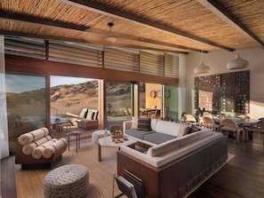 A spacious living area with a large sofa, a coffee table, and a dining table. There are multiple chairs around the dining table and a large window offering a view of the desert. - Six Senses Southern Dunes, The Red Sea (Umluj)