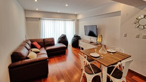 A modern living room with a brown leather sofa, a wooden dining table with chairs, a flat-screen TV mounted on the wall, and a large window with sheer curtains. - Beutifull 2 bedroom apartment in the center of Miraflores! Great Location (Miraflores)
