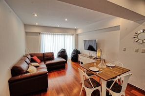 Uma sala de estar moderna com um sofá de couro marrom, uma mesa de jantar de madeira com cadeiras, uma TV de tela plana na parede e uma janela grande com cortinas leves.