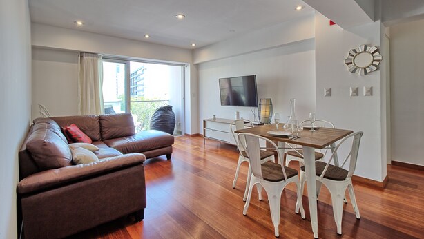 A modern living room with a brown leather sofa, a dining table with white chairs, a flat-screen TV mounted on the wall, and a large window with curtains. - Beutifull 2 bedroom apartment in the center of Miraflores! Great Location (Miraflores)