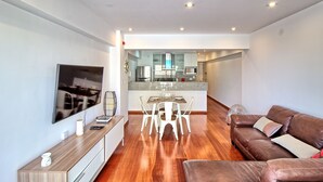 A modern living room with a brown leather sofa, a dining table with white chairs, a flat-screen TV mounted on the wall, and a kitchen area in the background. - Beutifull 2 bedroom apartment in the center of Miraflores! Great Location (Miraflores)