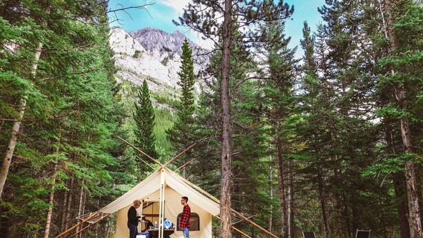 Una piccola tenda aperta con un tavolo da picnic e una zona notte, circondata da alti pini e una montagna sullo sfondo.