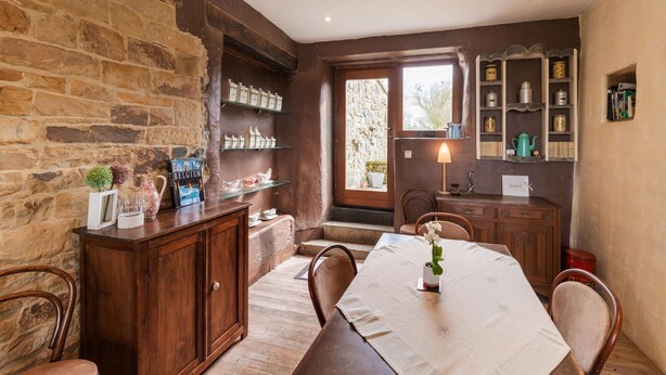 A cozy dining area with a wooden table set for two, surrounded by chairs. A stone wall and a wooden cabinet with shelves are visible in the background. - Tranquil Home in Tinlot (Tinlot)