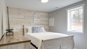Un dormitorio con cabecera de madera, cama con ropa de cama blanca, mesita de noche con lámpara y ventana con vistas al exterior.