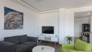 Un salon moderne avec un canapé gris, un fauteuil vert et une table basse blanche. Un téléviseur à écran plat est fixé au mur, et on aperçoit un coin cuisine à l’arrière-plan.