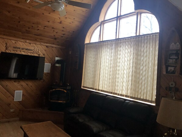 A cozy cabin interior with a fireplace, a television mounted on the wall, and a large window with curtains.