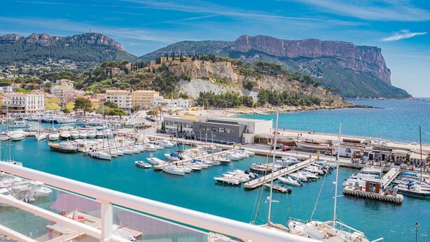 Una marina con numerosos barcos amarrados, un edificio con letrero y un paisaje montañoso al fondo.