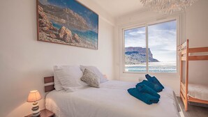 Un dormitorio con cama, mesita de noche, lámpara, un cuadro, ventana con vista al mar y una cama literera.