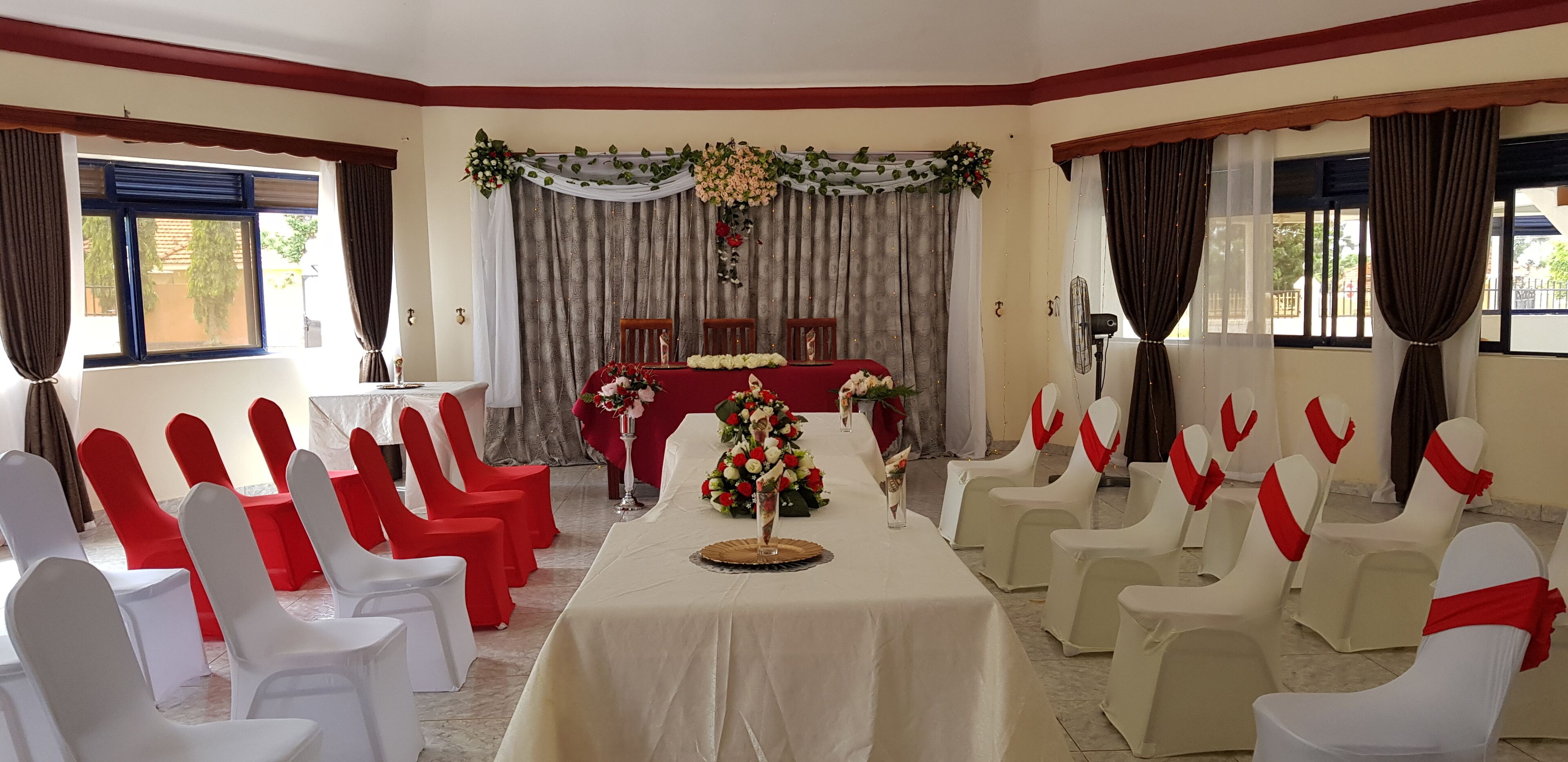 Un salón de banquetes con filas de sillas blancas y rojas, una mesa larga con centros de mesa florales y un fondo decorado con cortinas y adornos florales.